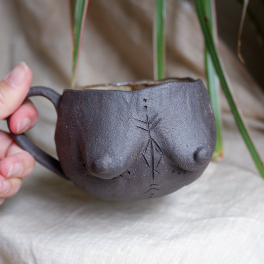 A handmade dark gray ceramic cup shaped like a stylized female breast, held by a hand against a soft beige fabric background with green plant leaves in the upper left.